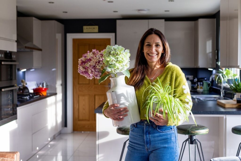 Smiling woman holding indoor plants in a modern kitchen filled with natural light